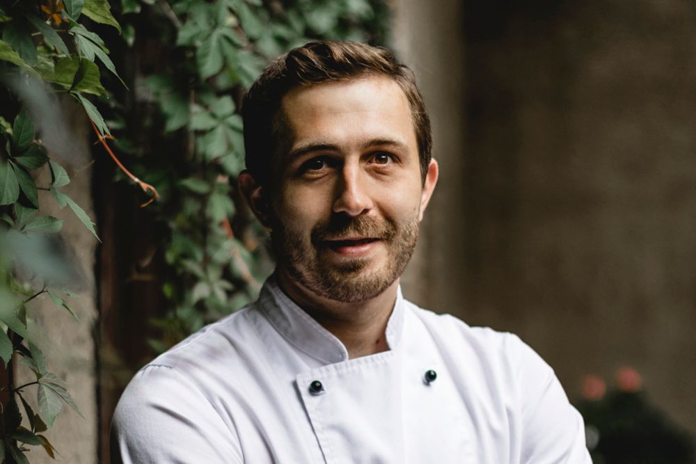 Ein Mann in weißer Kochjacke steht vor einer mit Efeu bewachsenen Wand.