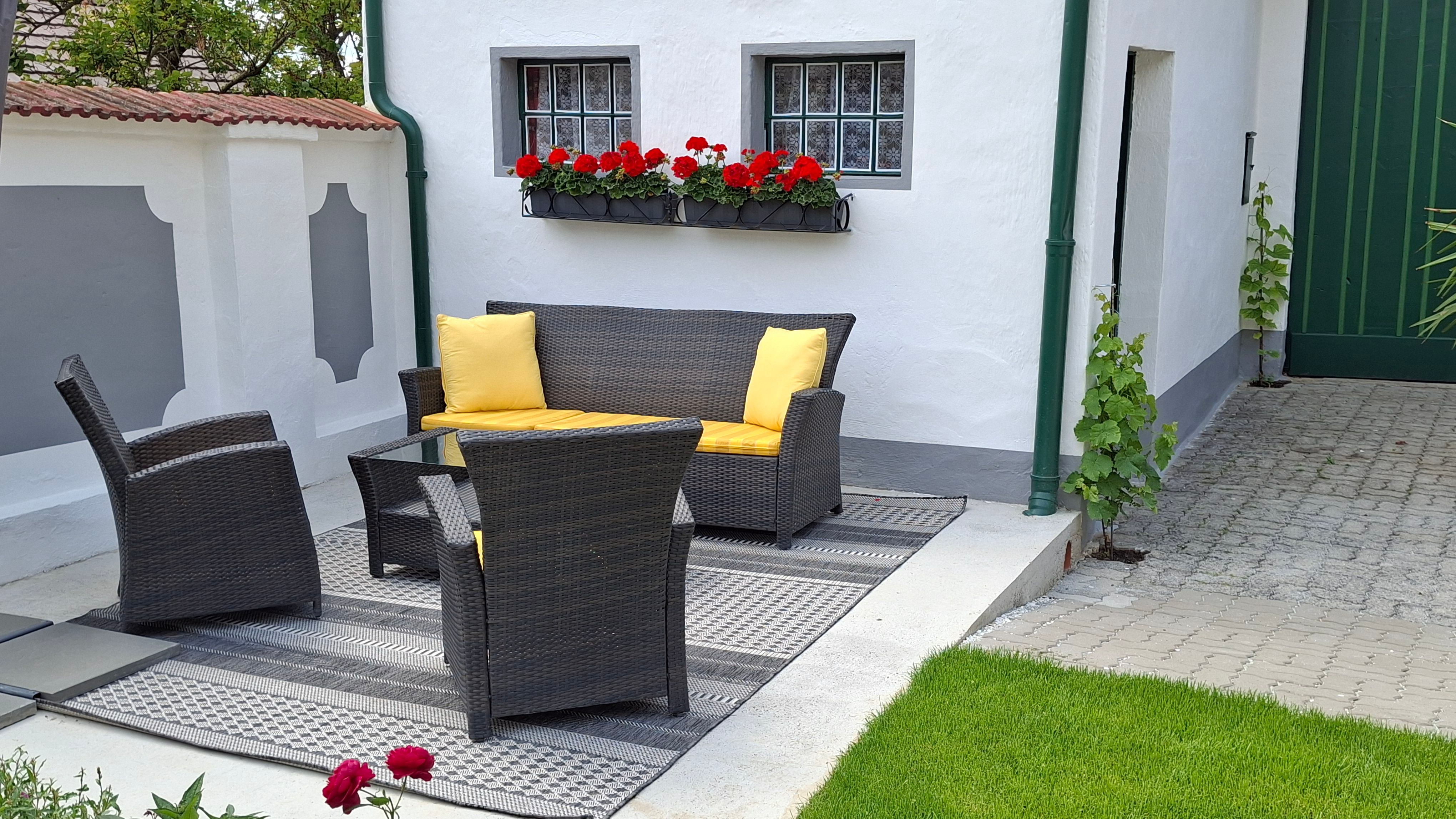 Terrasse mit Rattanmöbeln und gelben Kissen, umgeben von weißen Wänden und roten Blumen.