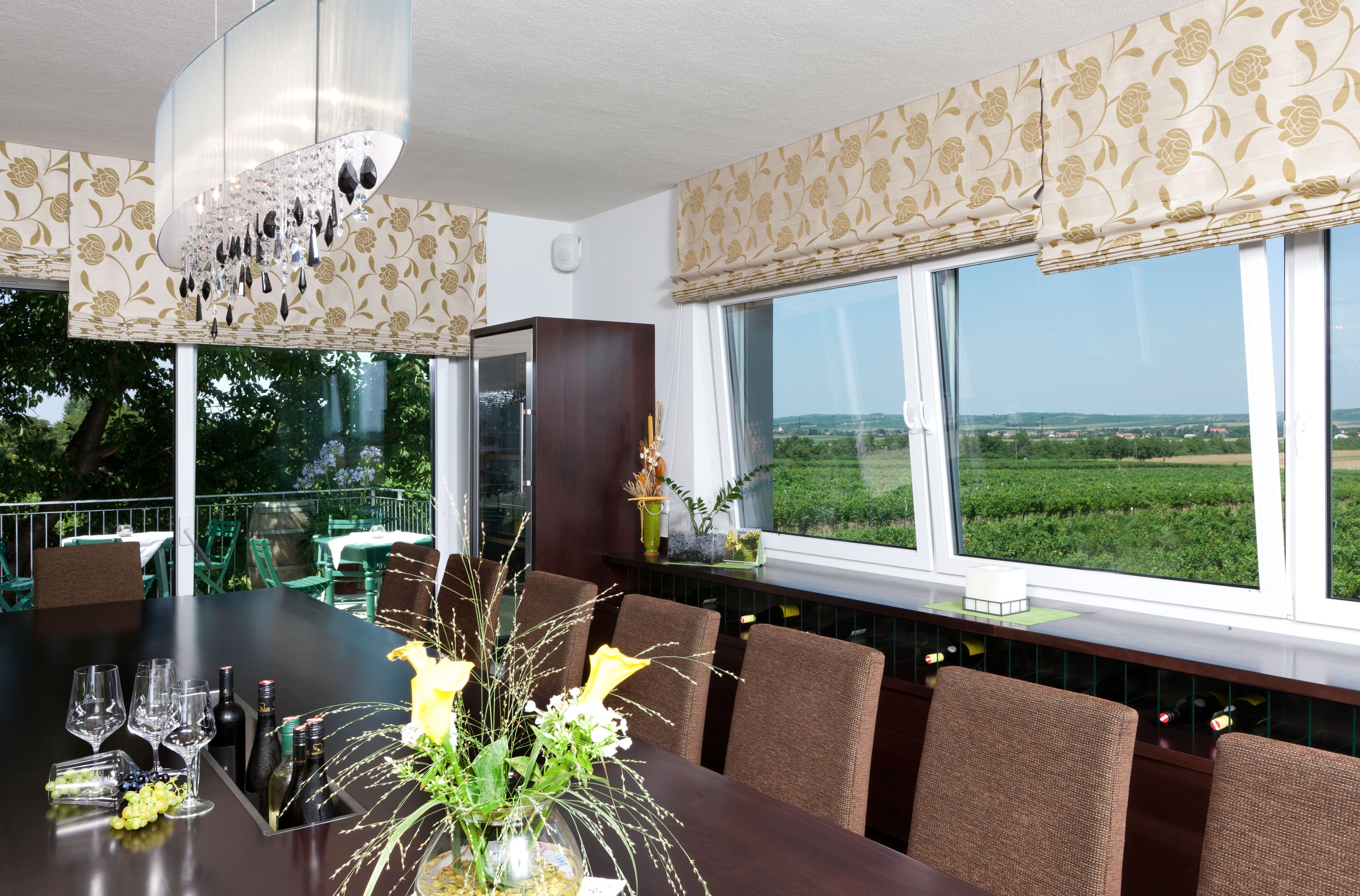 Ein Verkostungsraum mit einem großen Tisch, Weingläsern und Flaschen. Große Fenster bieten Blick auf Weinberge. Elegante Dekoration und Beleuchtung.