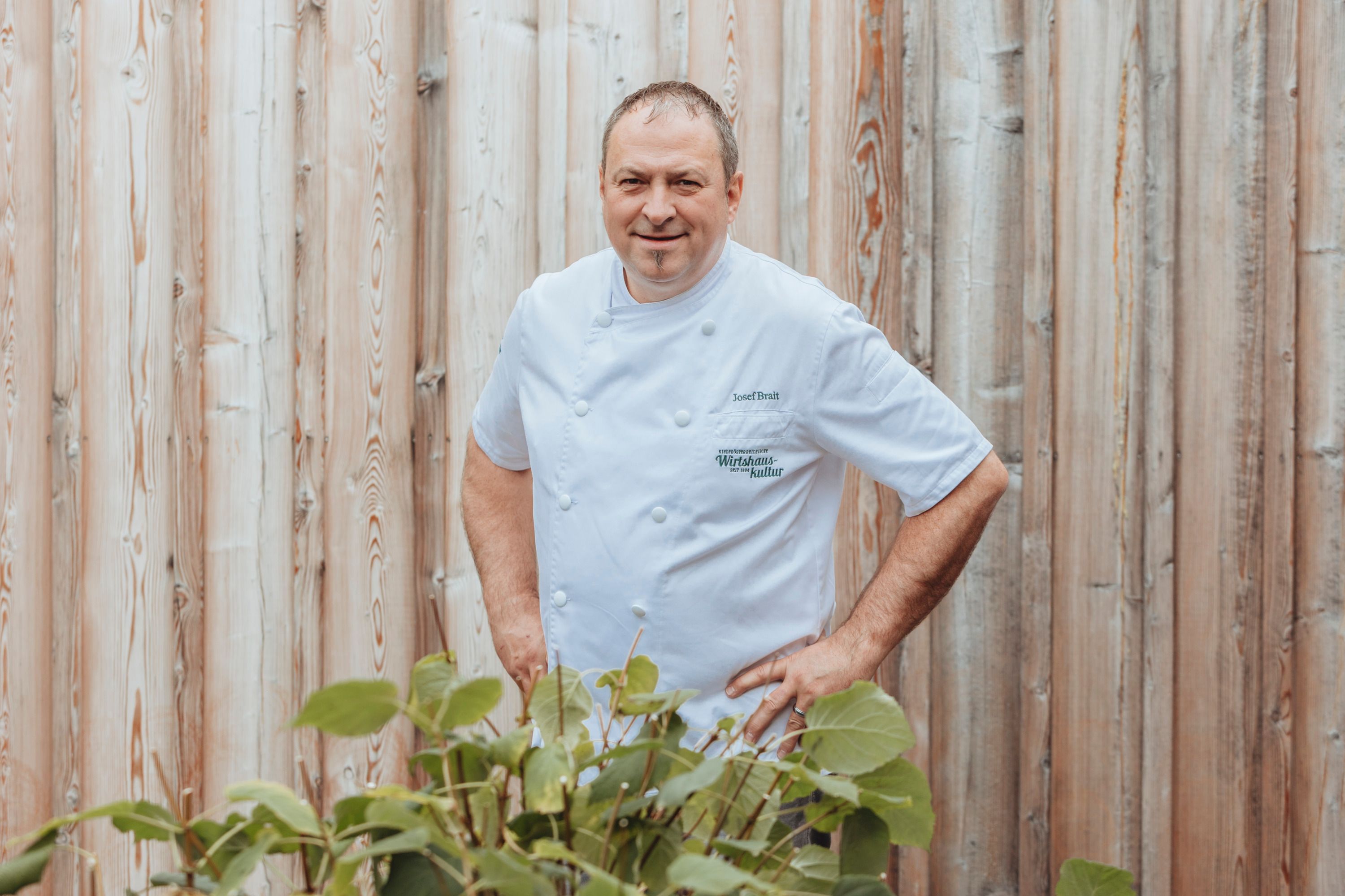 Ein Mann in weißer Kochjacke steht vor einer Holzwand.