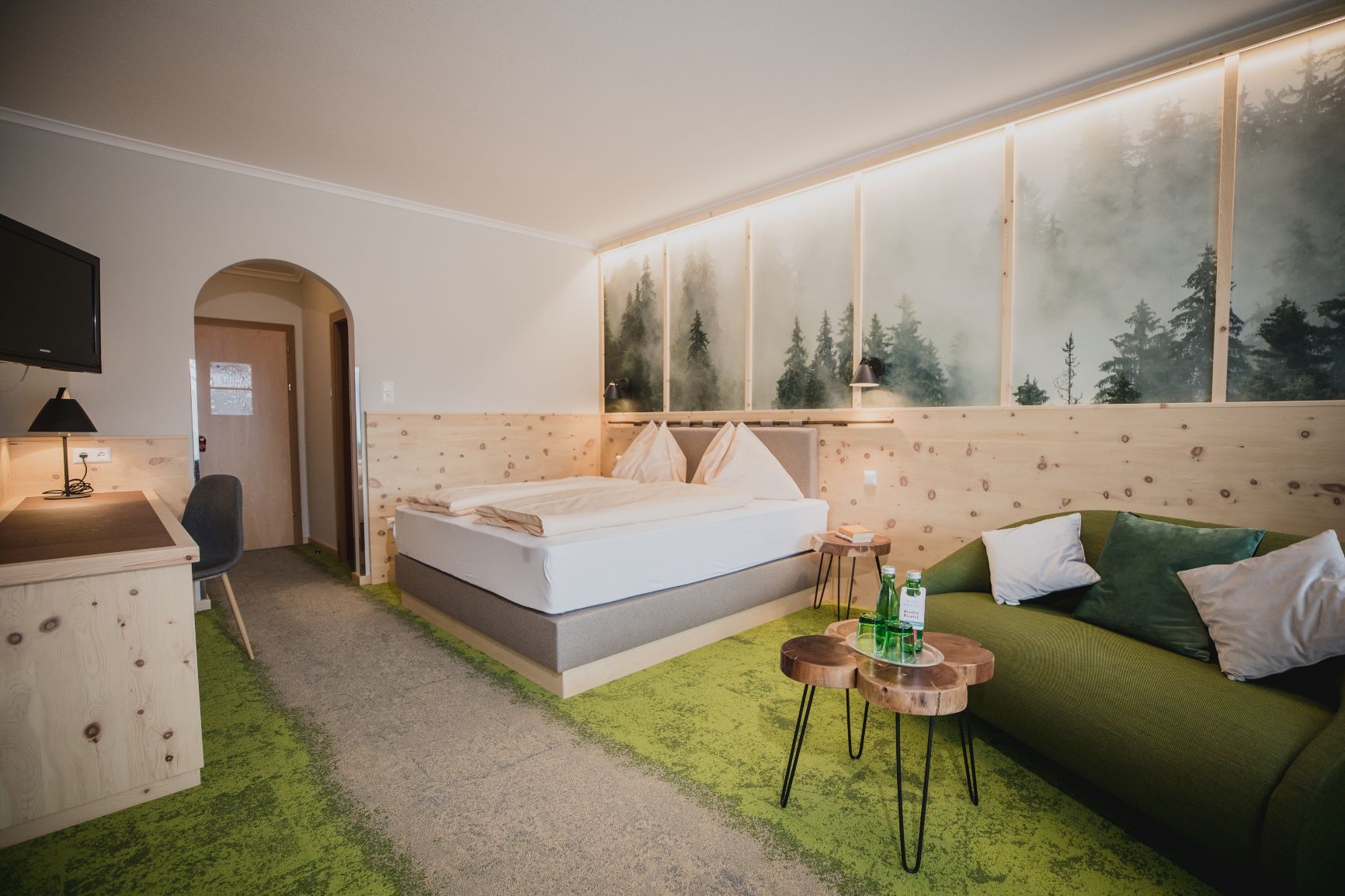 Ein gemütliches Hotelzimmer mit Zirbenholzmöbeln, einem Doppelbett, grünem Teppich und einem Sofa. An der Wand ein Bild von nebligem Wald.