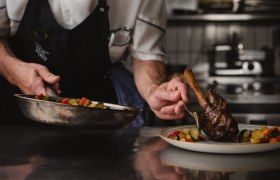 Ein Koch in einer professionellen Küche richtet ein Gericht mit Fleisch und Gemüse auf einem Teller an.