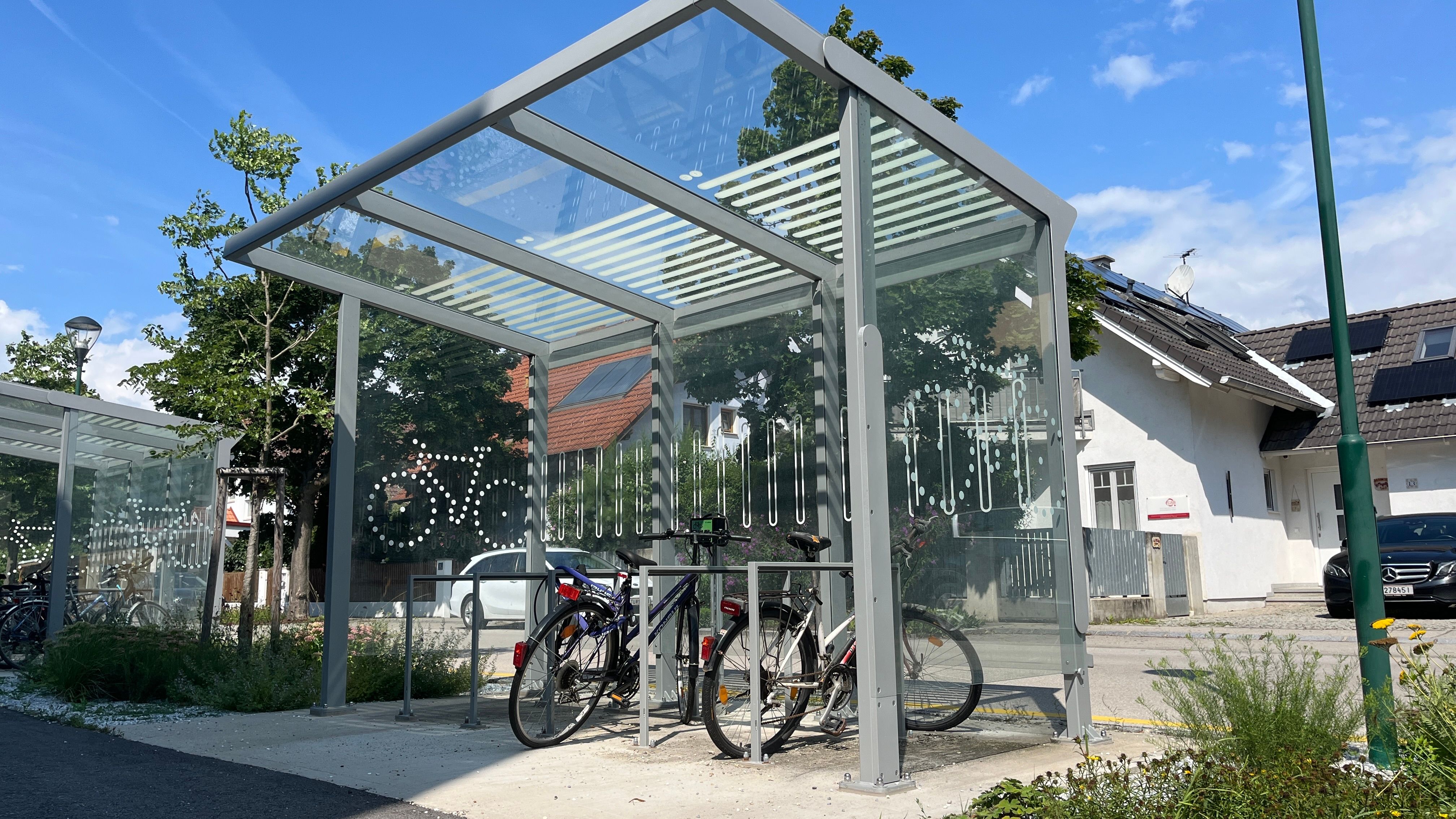 E-Bike-Ladestation mit überdachtem Bereich und Fahrrädern, im Hintergrund ein Haus mit Solarpanelen.