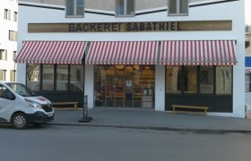 Au&szlig;enansicht der B&auml;ckerei Sabathiel mit rot-wei&szlig; gestreifter Markise und Lieferwagen davor.