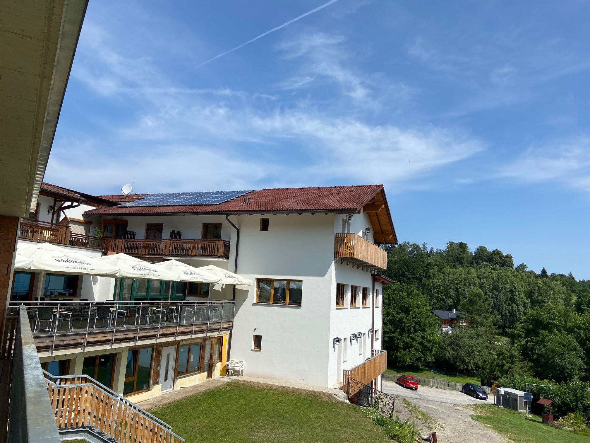 Blick auf ein Gebäude mit Terrasse und Sonnenschirmen, umgeben von grüner Landschaft und blauem Himmel.
