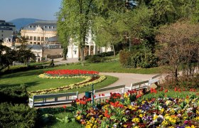 Kurpark Baden mit blühenden Blumenbeeten, Bänken und einem historischen Gebäude im Hintergrund.