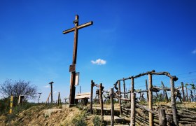 Holzkreuz auf einem Hügel mit Weinreben und blauem Himmel im Hintergrund.
