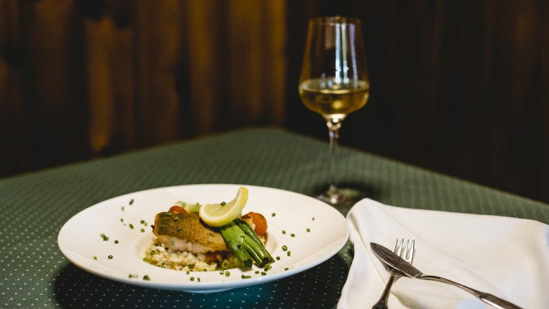 Ein Teller mit Lachsforelle und Risotto, garniert mit Zitrone und Gem&uuml;se, steht auf einem gedeckten Tisch neben einem Glas Wei&szlig;wein.