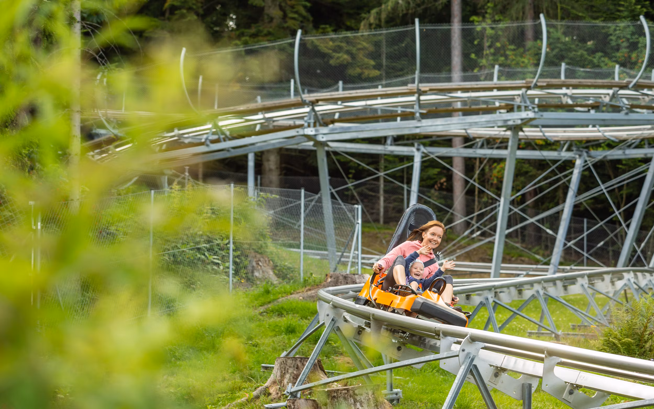 Eine Frau und ein Kind fahren lachend auf einer Sommerrodelbahn durch eine grüne Landschaft.