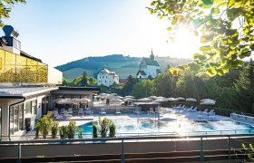 Außengelände eines Hotels mit Pool, Sonnenschirmen und Liegen, umgeben von Natur und Kirche im Hintergrund.