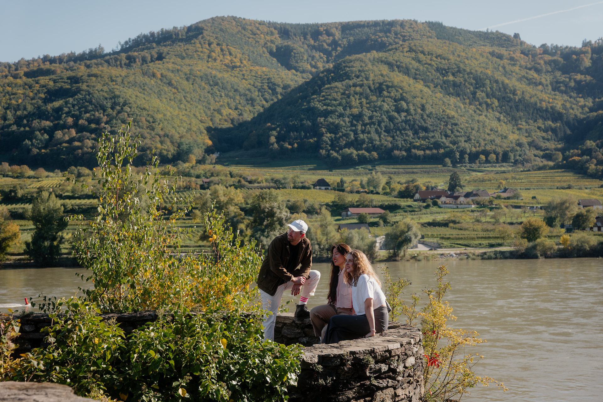 Drei Personen, teilweise sitzend auf einer niedrigen Steinmauer am Ufer der Donau; im Hintergrund breitet sich eine grüne Hügellandschaft mit Feldern und Wald aus.