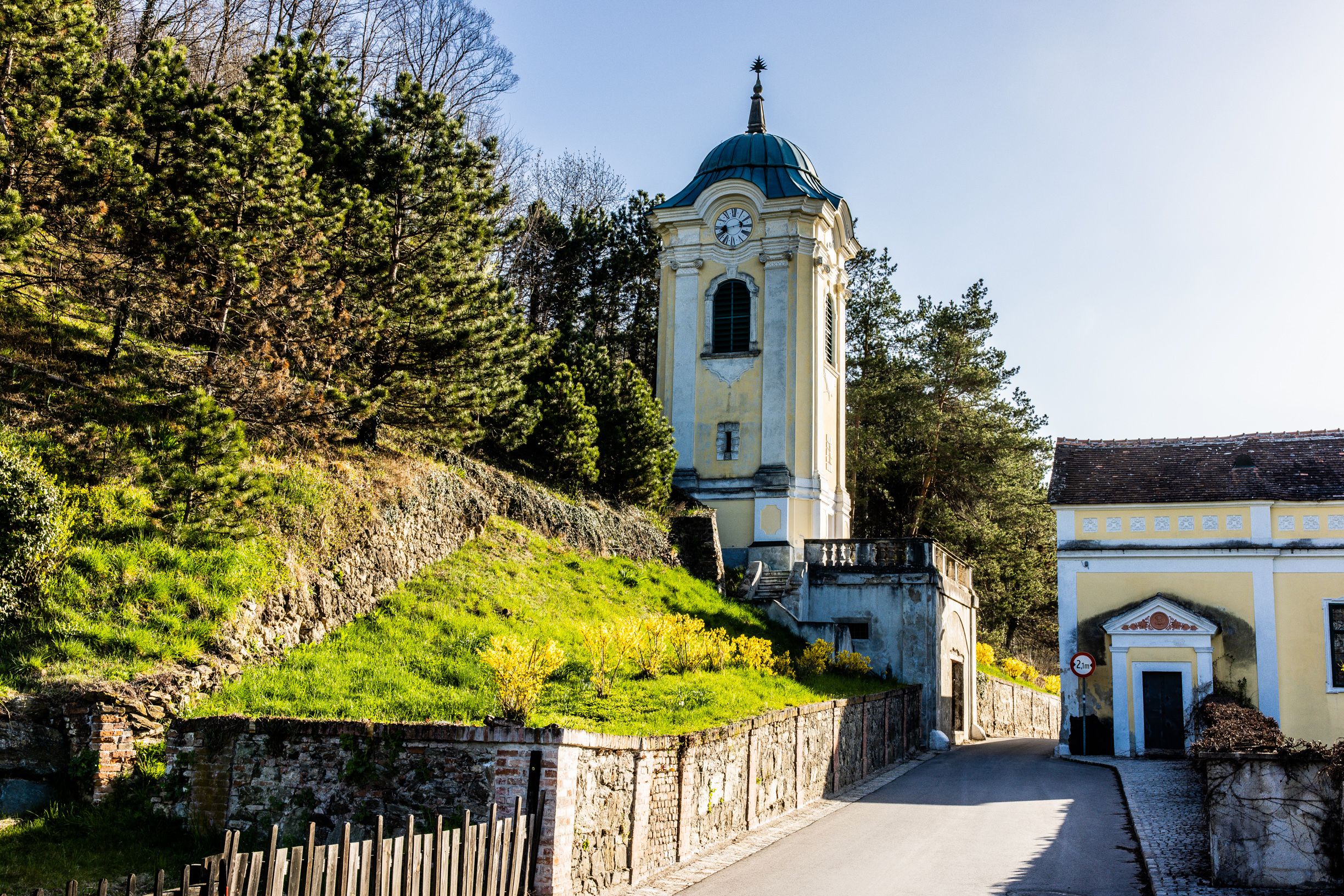 Eine gelber Uhrturm in Bad Erlach, umgeben von Bäumen und einem sonnigen Hang, eine schmale Straße führt vorbei.