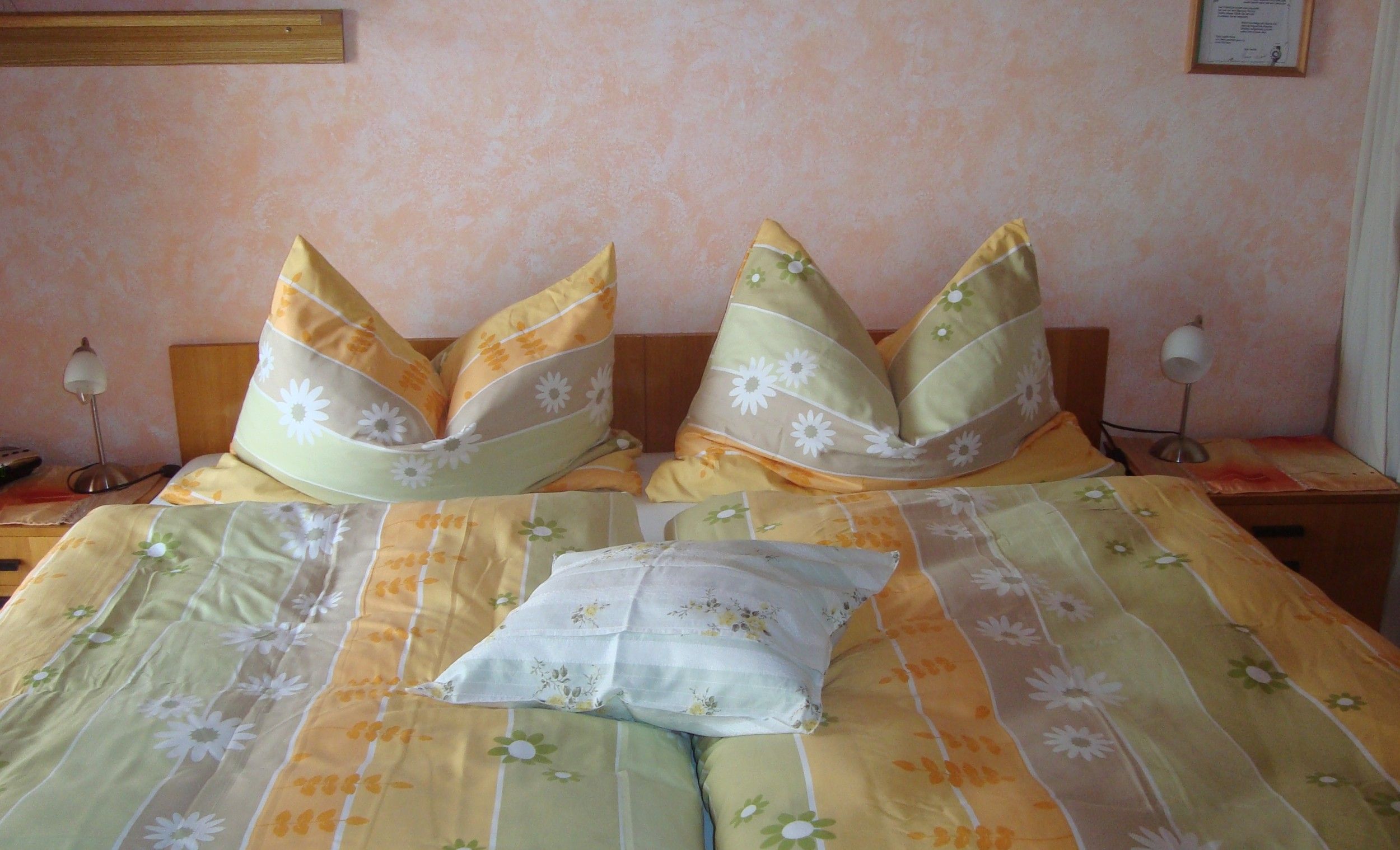 Ein gemütliches Schlafzimmer mit einem Doppelbett, das mit gelb-grüner Bettwäsche und Blumendekor bezogen ist. Zwei Nachttischlampen stehen daneben.