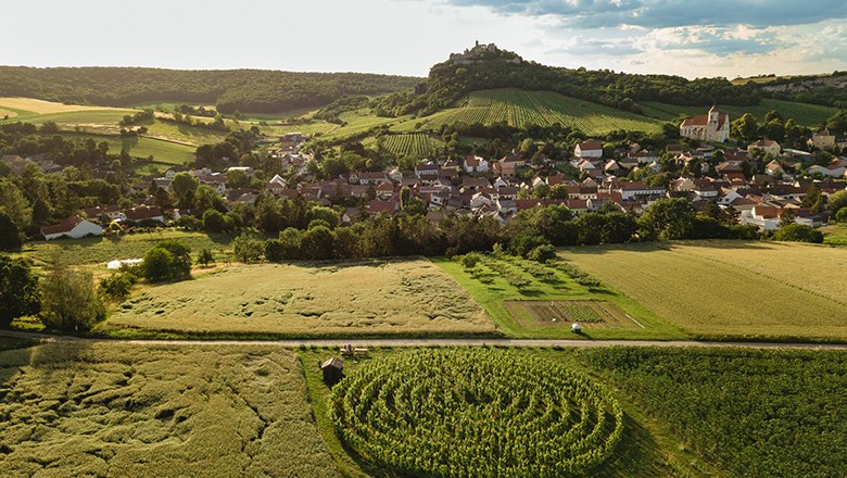 Luftaufnahme eines Dorfes mit Feldern und einer Burg auf einem H&uuml;gel im Hintergrund.