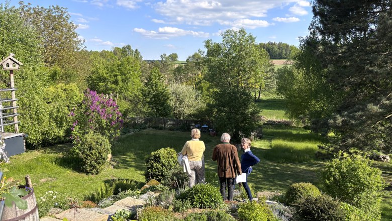 Drei Personen stehen in einem grünen Garten mit Bäumen und Sträuchern, umgeben von einer ländlichen Landschaft unter blauem Himmel mit Wolken.