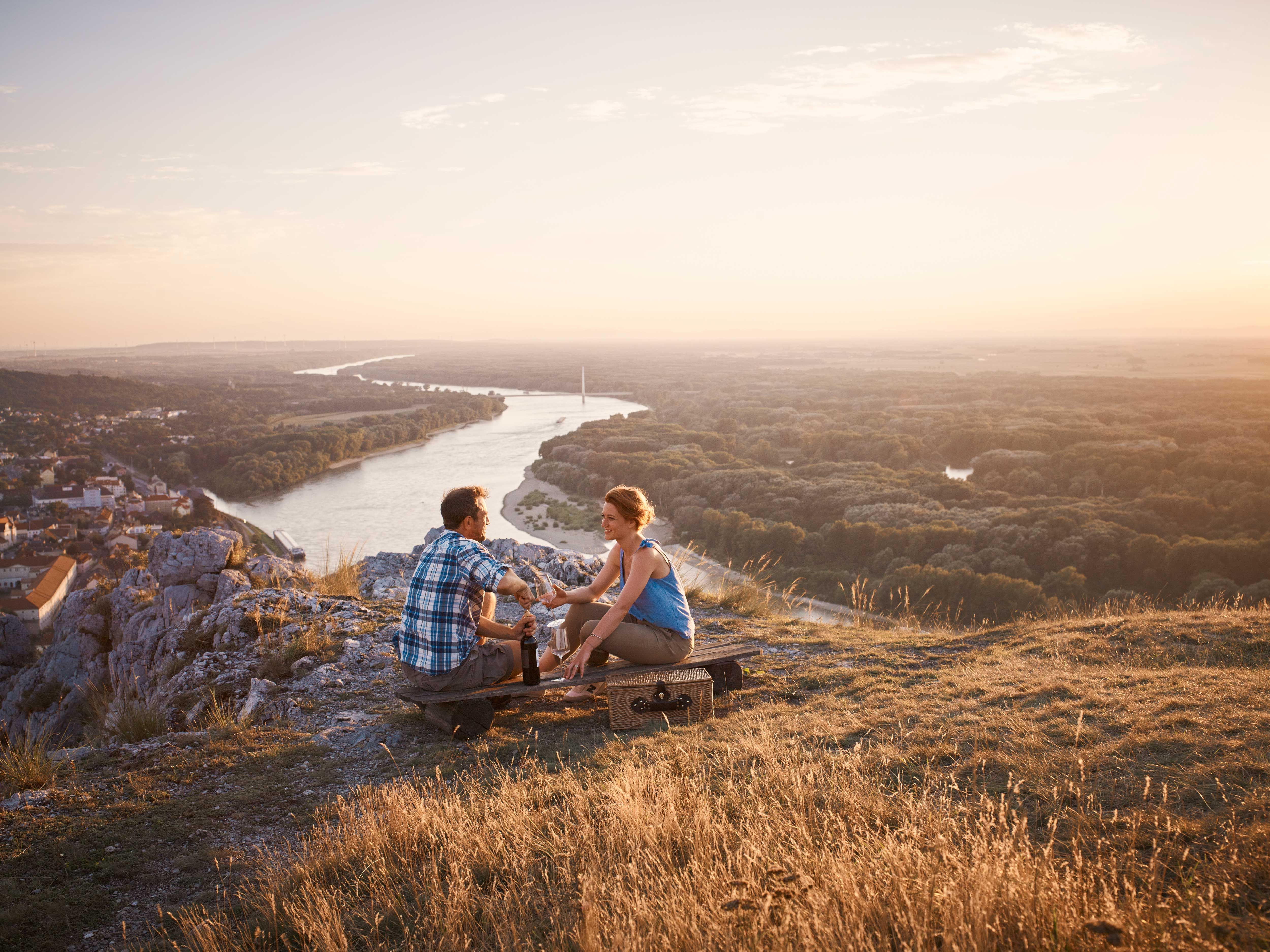 Zwei Personen sitzen auf einem Hügel mit Blick auf einen Fluss und eine Stadt im Sonnenuntergang.