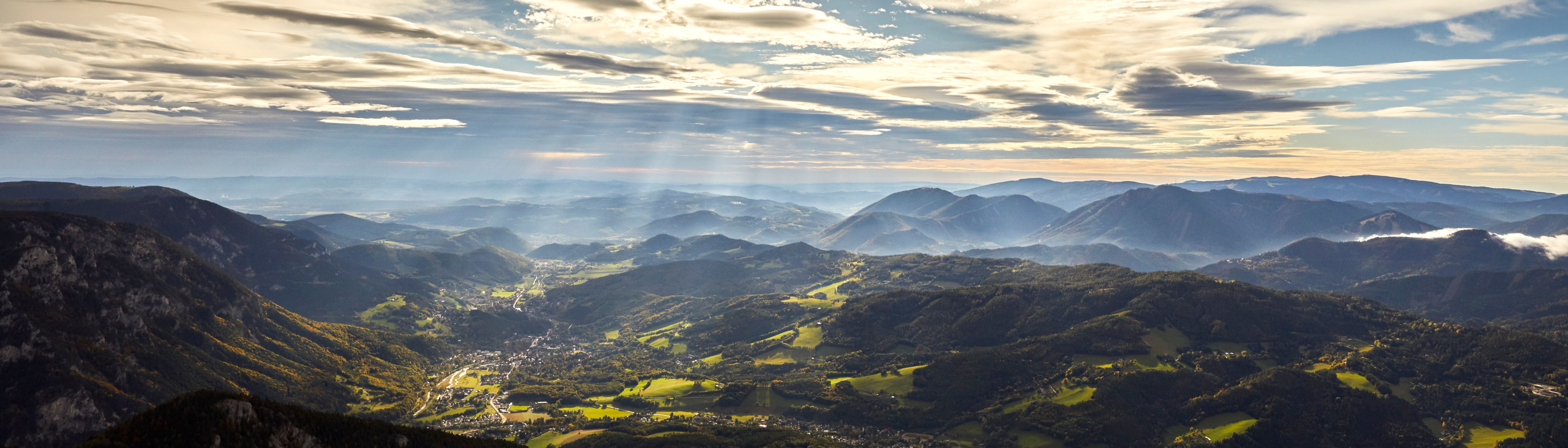 Panoramablick auf die Rax-Alpen mit bewaldeten Hügeln und Sonnenstrahlen durch Wolken.