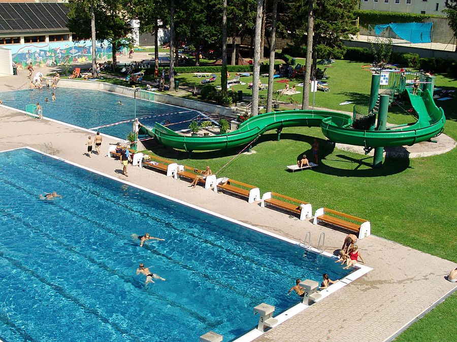 Ein Freibad mit zwei Schwimmbecken, einer grünen Wasserrutsche und Liegewiese.