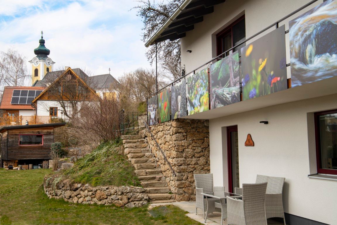 Ein modernes Haus mit Balkon und Naturbildern, daneben eine Kirche mit Zwiebelturm und ein weiteres Gebäude mit Solarpanelen.