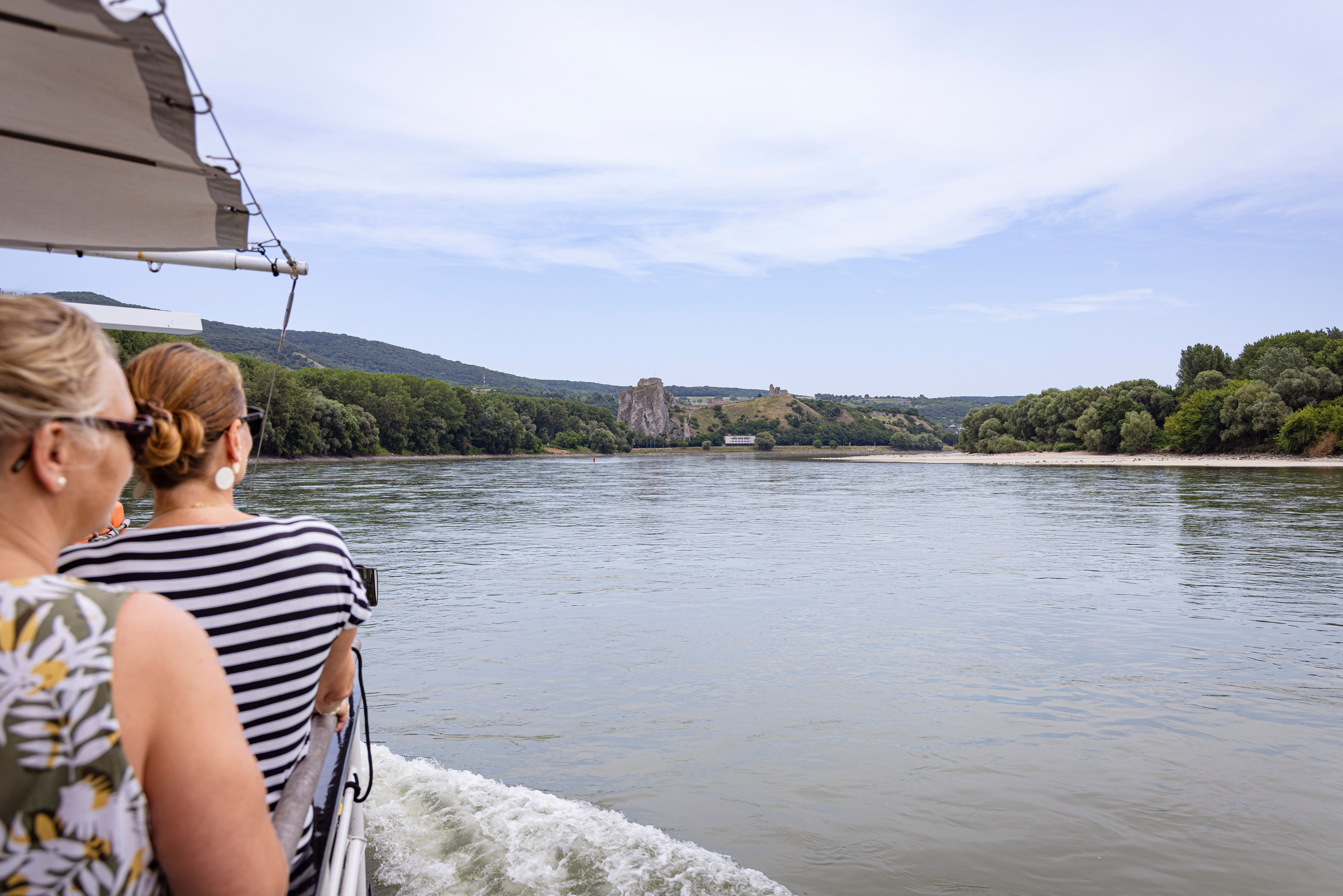 Zwei Frauen auf einem Boot blicken auf eine Flusslandschaft mit Hügeln und Ruinen.