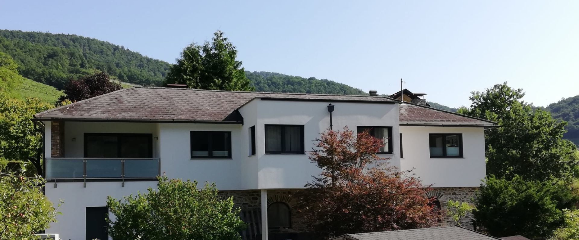 Außenansicht eines modernen weißen Ferienhauses mit Bäumen im Vordergrund und bewaldeten Hügeln im Hintergrund.