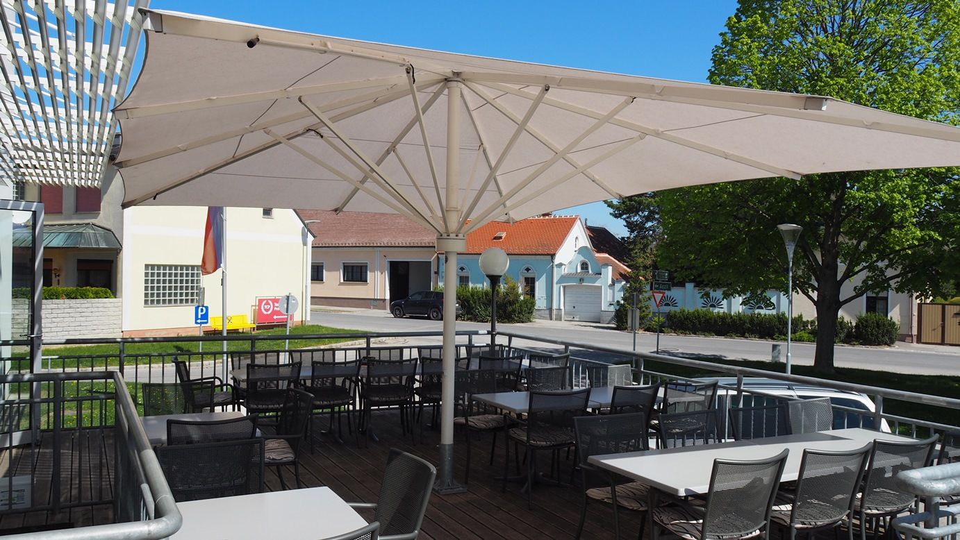 Terrasse eines Restaurants mit Sonnenschirm und leeren Tischen und Stühlen, im Hintergrund eine Straße und Gebäude.