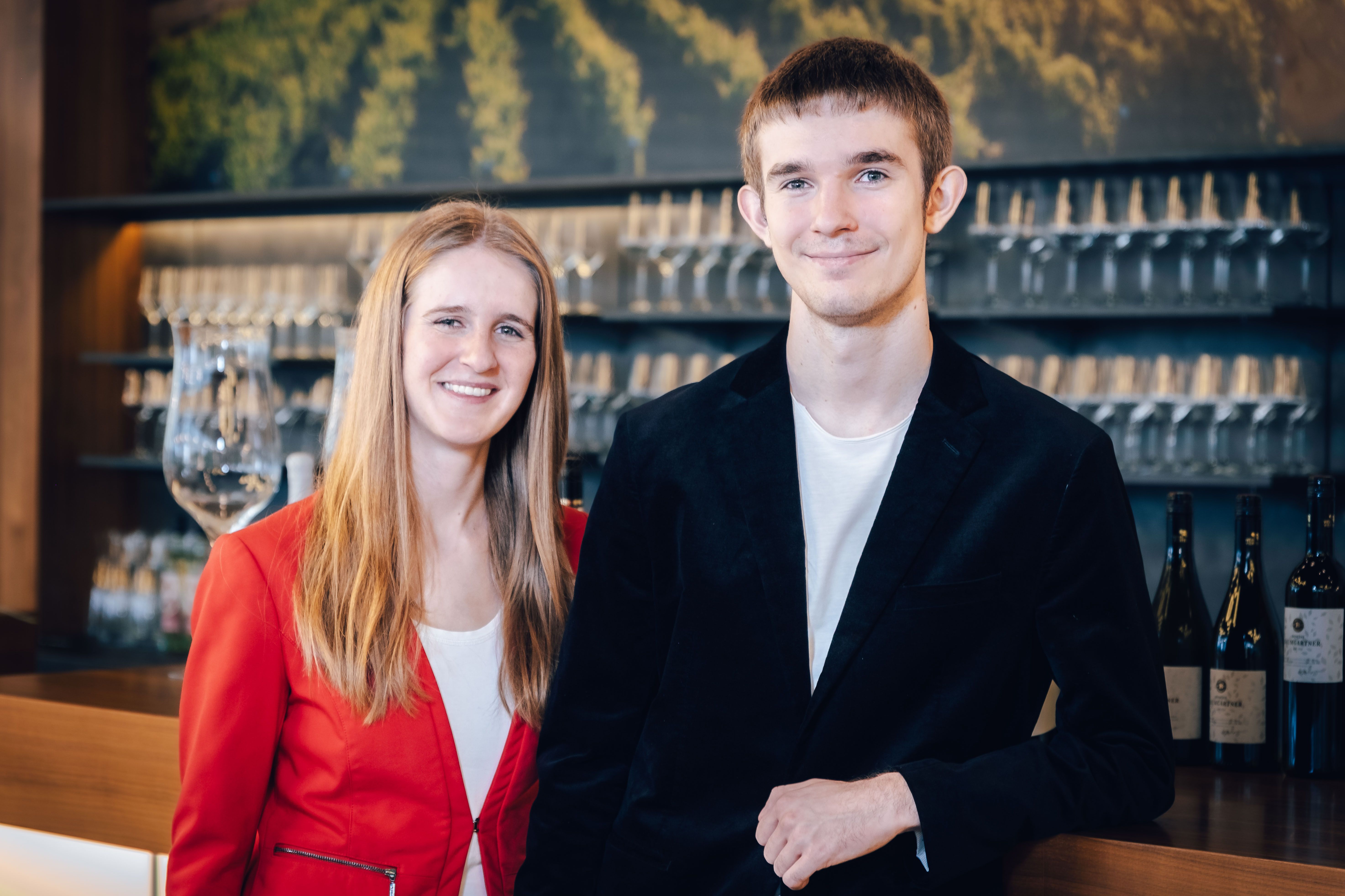 Katharina und Lorenz Baumgartner in eleganter Kleidung vor einem Regal mit Weingläsern.