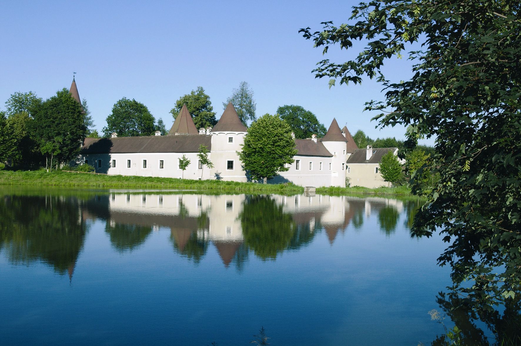 Schloss mit spitzen Türmen spiegelt sich in einem ruhigen See, umgeben von Bäumen.