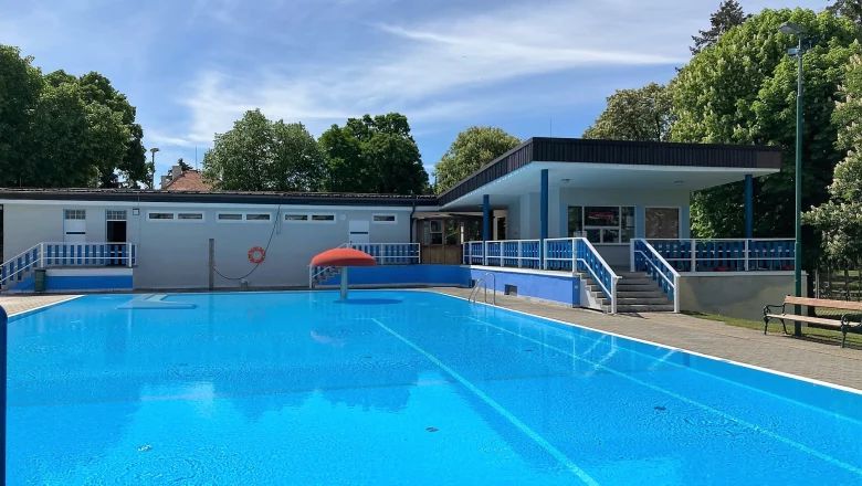 Ein leeres Freibad mit blauem Wasser und einem roten Pilzbrunnen, umgeben von Bäumen und einem Gebäude im Hintergrund.
