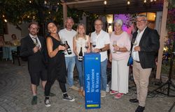 Sieben Personen stehen bei einer Abendveranstaltung im Freien mit Weingläsern und Weinflaschen rund um eine Stele mit der Aufschrift „Kultur bei Winzerinnen & Winzern“