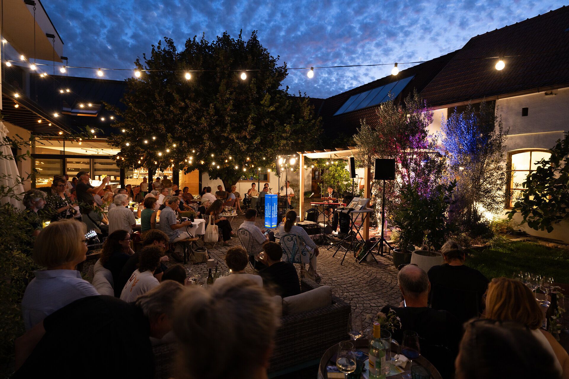 Abendliches Konzert in einem stimmungsvoll beleuchteten Innenhof: Zahlreiche Gäste sitzen an Tischen und verfolgen den Auftritt eines Musikers.