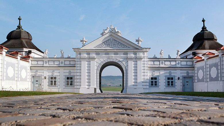 Barockes Tor mit weißen Verzierungen und Figuren, gepflasterter Weg im Vordergrund.
