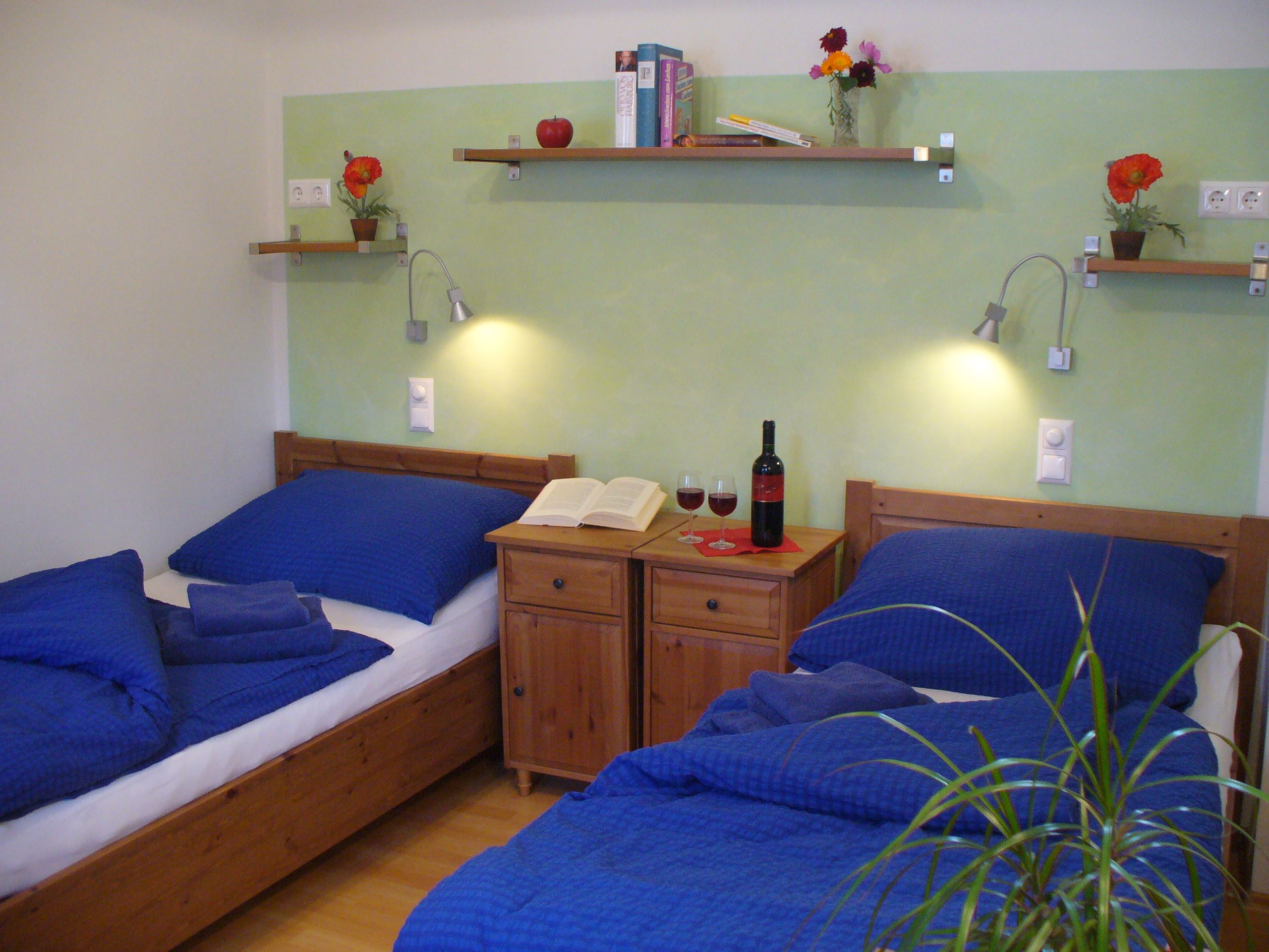 Zweibettzimmer mit blauer Bettwäsche, Nachttisch mit Weinflasche und Gläsern, Bücherregale und Pflanzen.