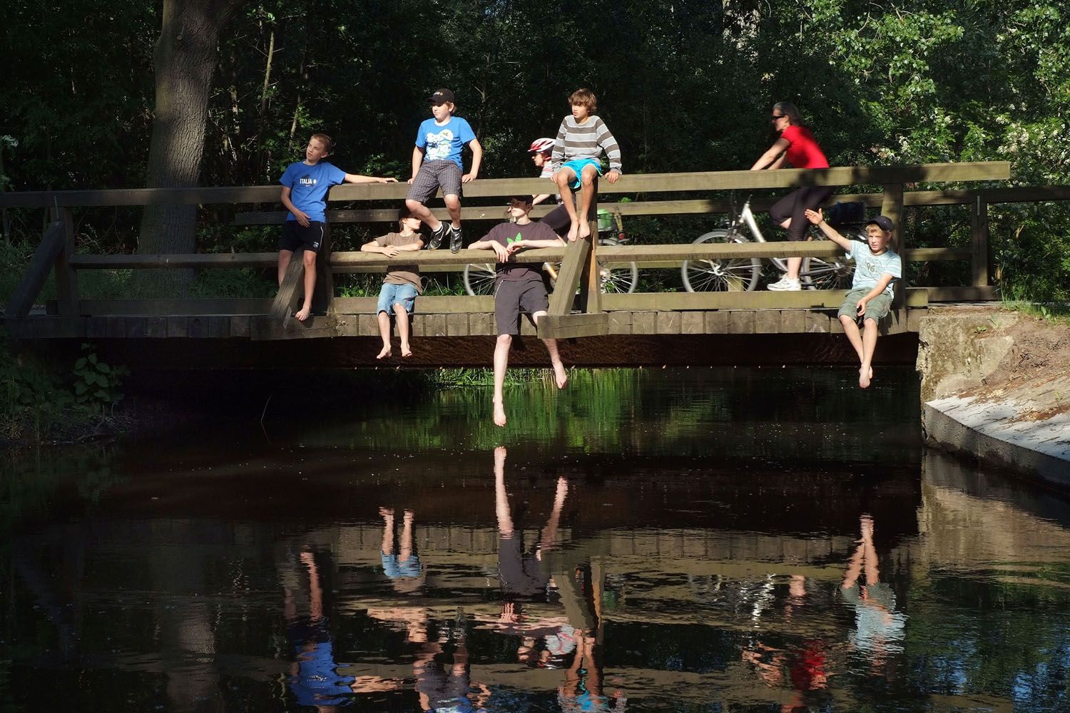 Menschen sitzen auf einer Holzbrücke über einem Fluss, einige lassen die Beine baumeln. Eine Person fährt mit dem Fahrrad über die Brücke.