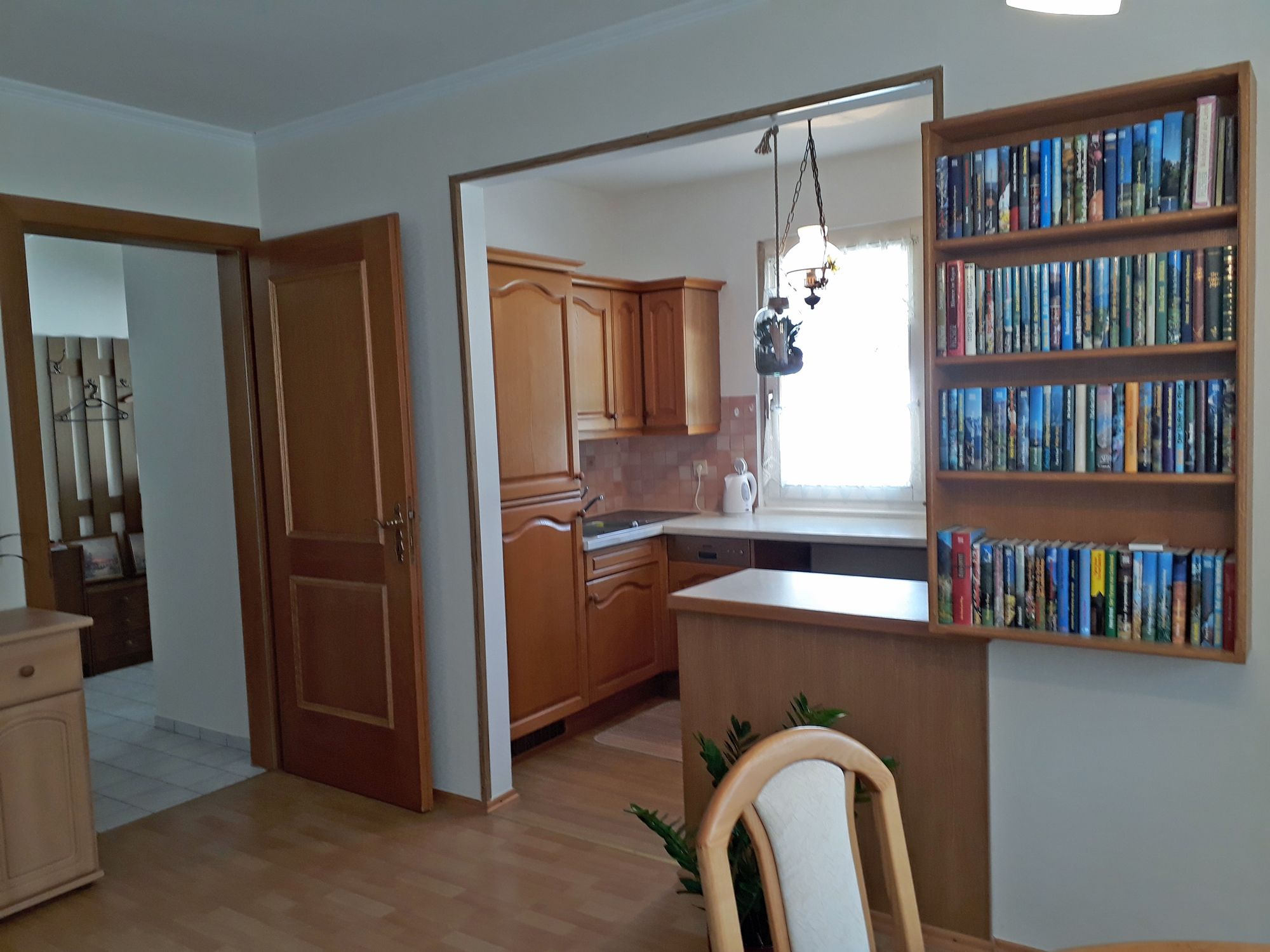 Innenansicht einer Küche mit Holzschränken, einem Bücherregal und einem Esstisch.