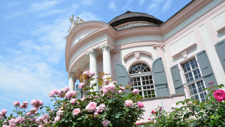 Barocker Pavillon mit rosa Fassade und blühenden Rosen im Vordergrund.