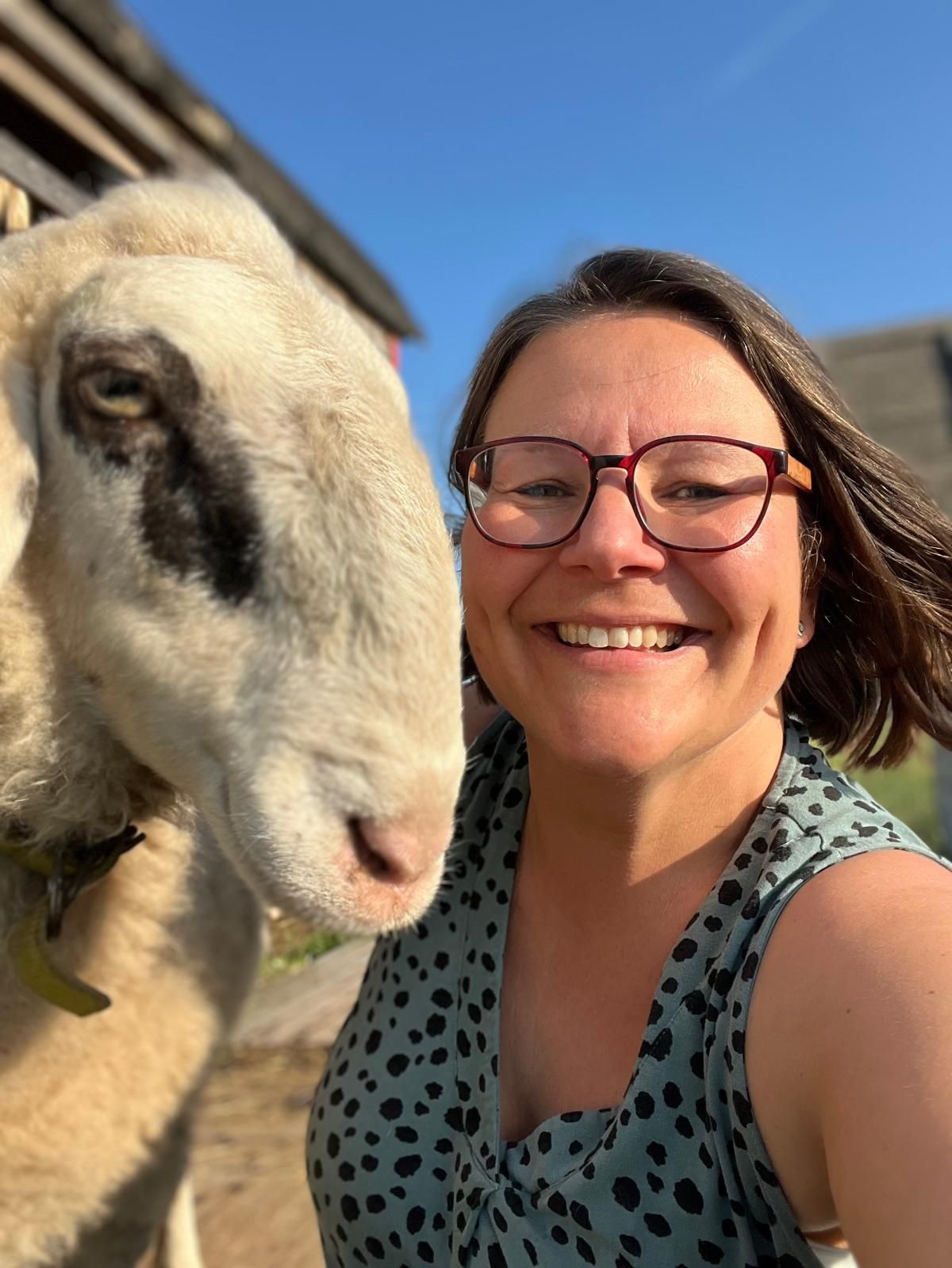 Frau mit Brille lächelt neben einem Schaf in der Sonne.