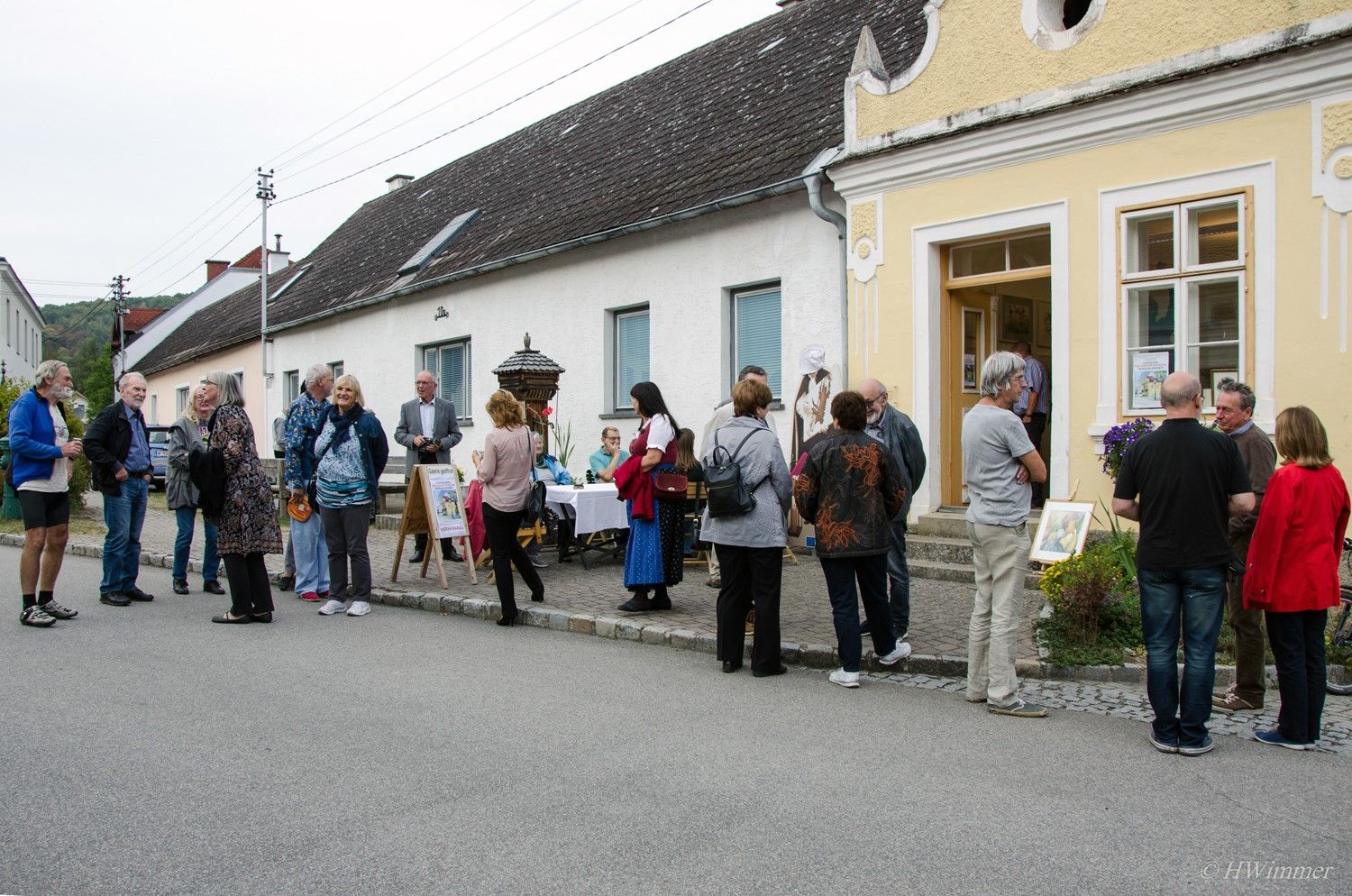 Eine Gruppe von Menschen steht vor einem gelben Gebäude auf einer Straße und unterhält sich. Einige betrachten Kunstwerke, die draußen ausgestellt sind.
