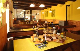 Innenansicht eines gem&uuml;tlichen Heurigen mit Holztischen, gelben Tischdecken und einer Auswahl an K&auml;se, Brot und Wein auf einem Tisch.