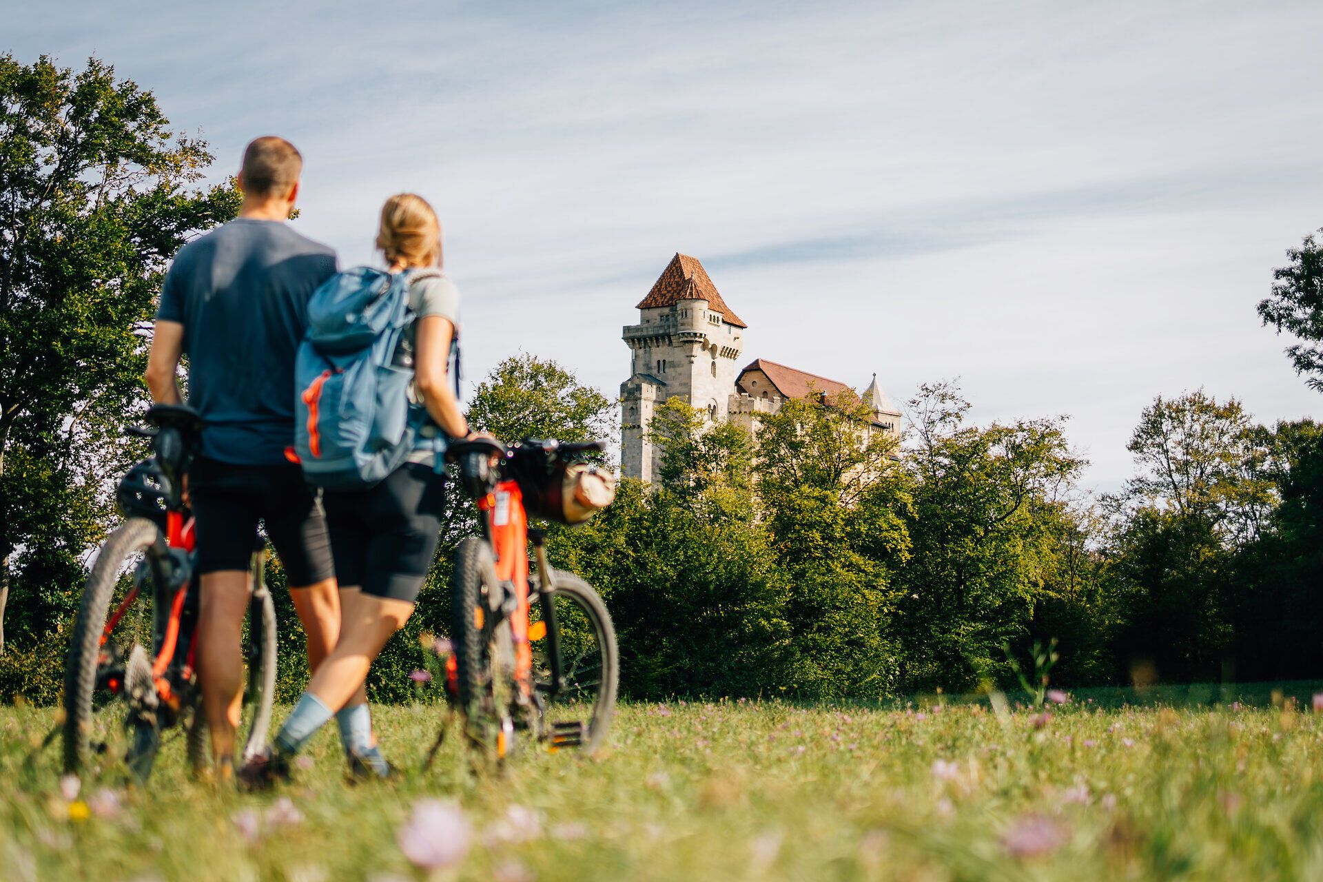 Ein Paar genießt die frische Luft und die malerische Aussicht auf die majestätische Burg Liechtenstein, während sie mit ihren Fahrrädern durch die sanften Wiesen des Wienerwaldes radeln. Die sanften Hügel und das satte Grün der Natur schaffen eine perfekte Kulisse für ein unvergessliches Abenteuer in der Natur.