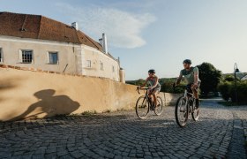 Zwei Radfahrer fahren auf einer gepflasterten Stra&szlig;e entlang einer alten Stadtmauer in Drosendorf.