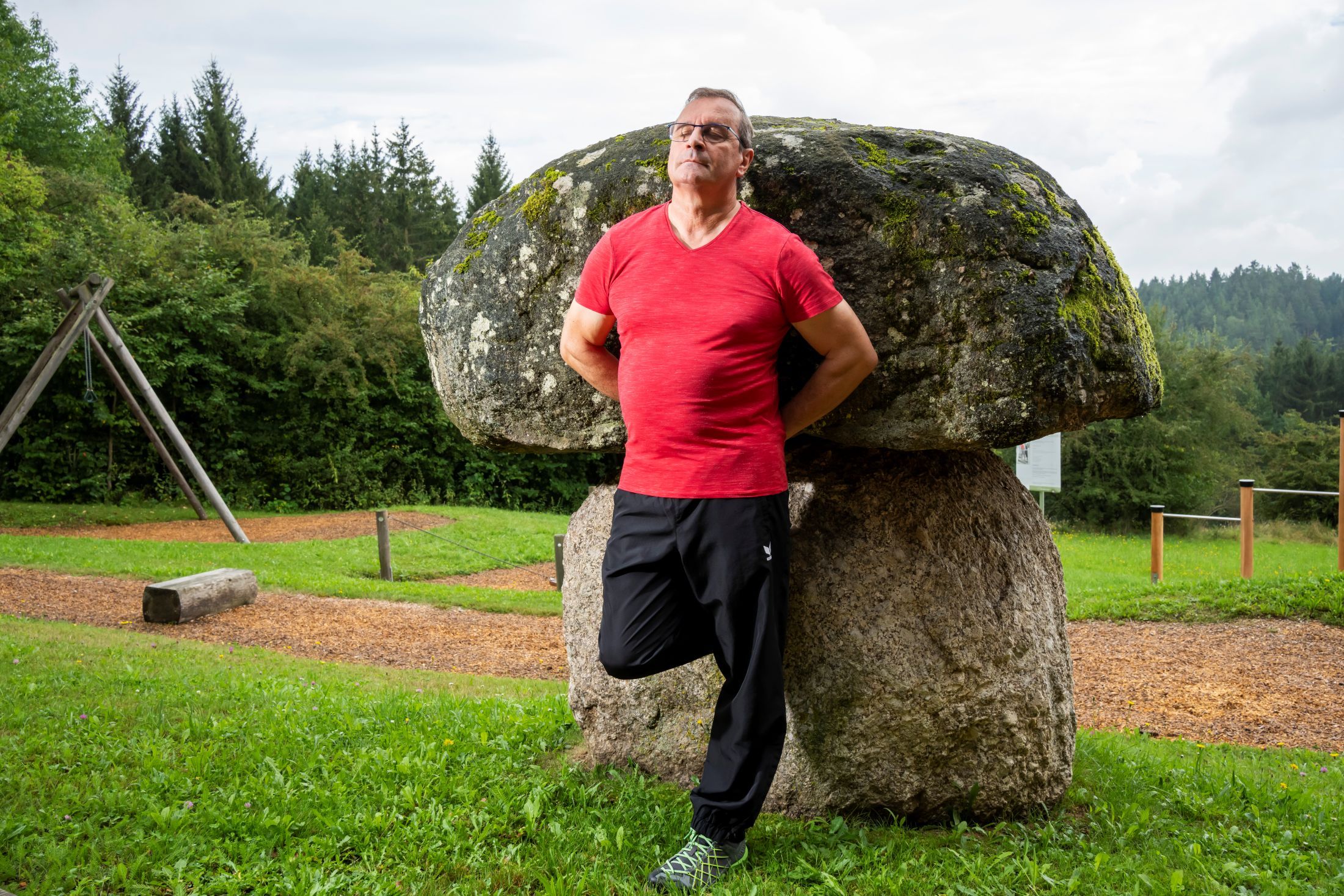 Ein Mann in rotem T-Shirt lehnt sich an einen großen, moosbedeckten Stein im Freien.