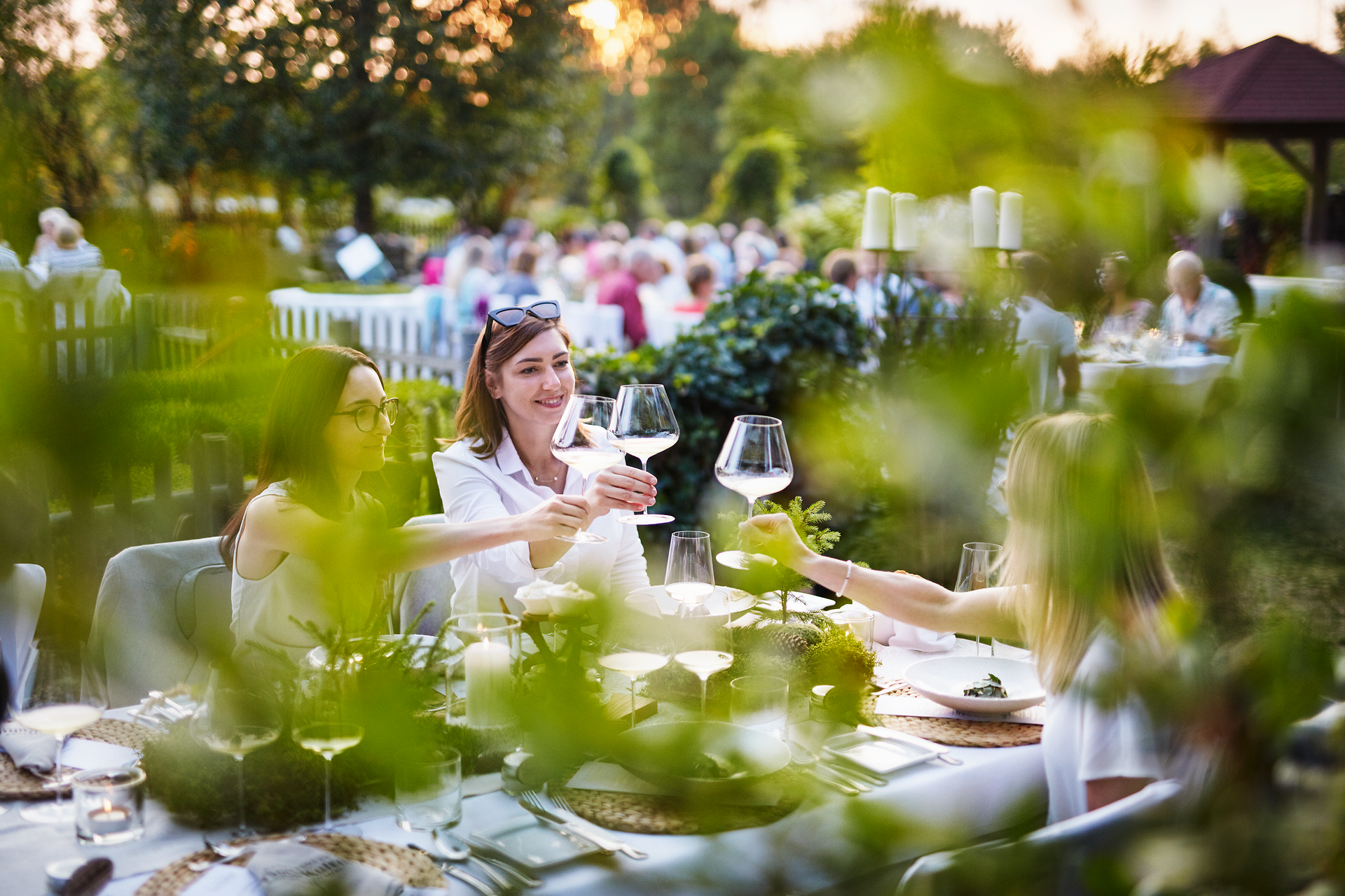 Umgeben von üppigem Grün und der warmen Abendsonne, genießen die Gäste ein geselliges Beisammensein. Die Tische sind liebevoll gedeckt und laden dazu ein, regionale Köstlichkeiten zu probieren und den Sommer in vollen Zügen zu erleben.