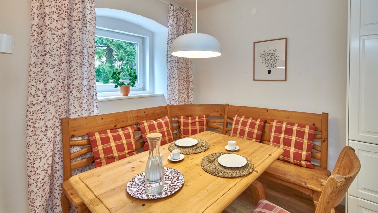 Gem&uuml;tlicher Essbereich mit Holztisch, Eckbank und karierten Kissen.