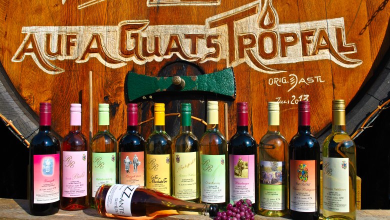 Verschiedene Weinflaschen vor einem gro&szlig;en Holzfass mit der Aufschrift 'Auf a guats Tropfal'.