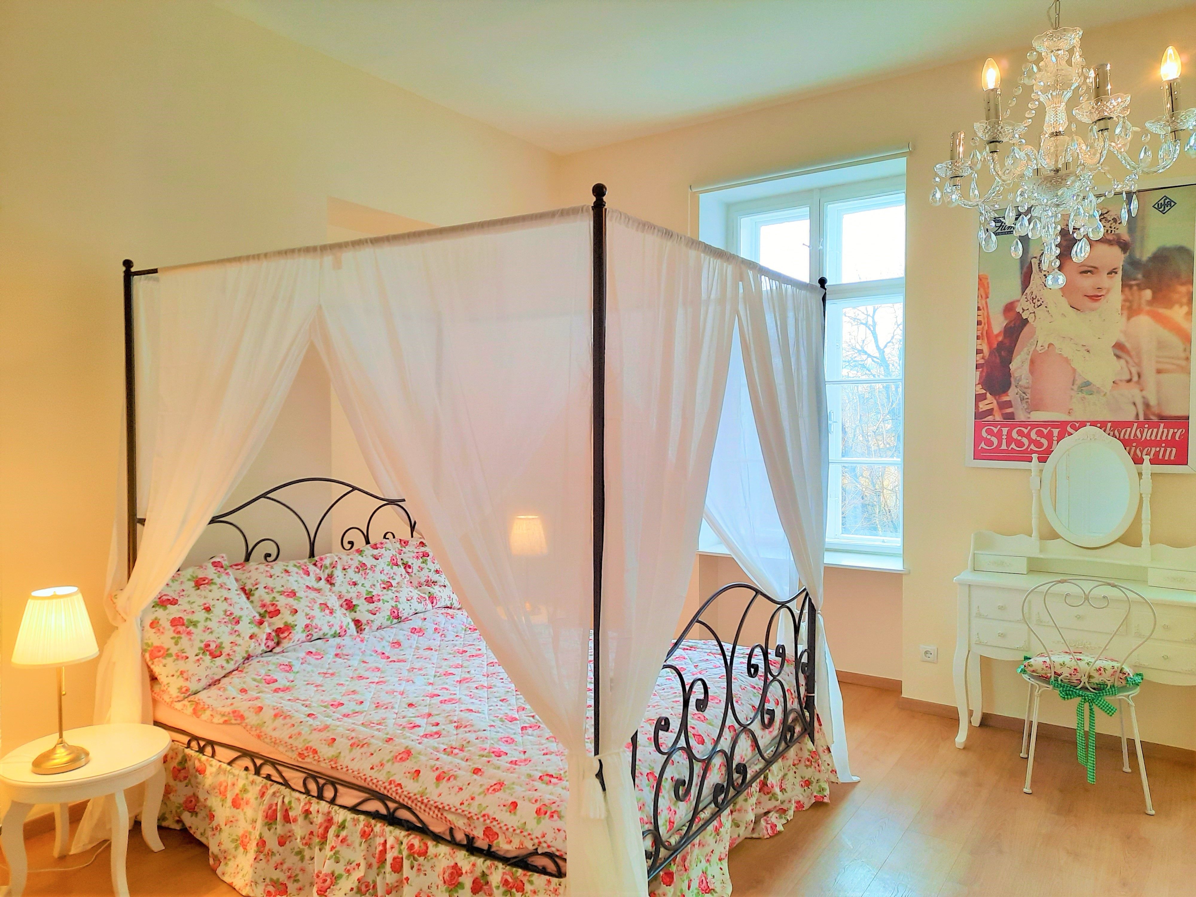 Schlafzimmer mit Himmelbett, Blumenbettwäsche, Kronleuchter und Schminktisch.