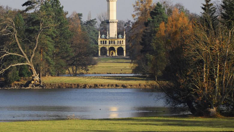 Ein hoher Turm im Hintergrund, umgeben von B&auml;umen, mit einem See im Vordergrund.