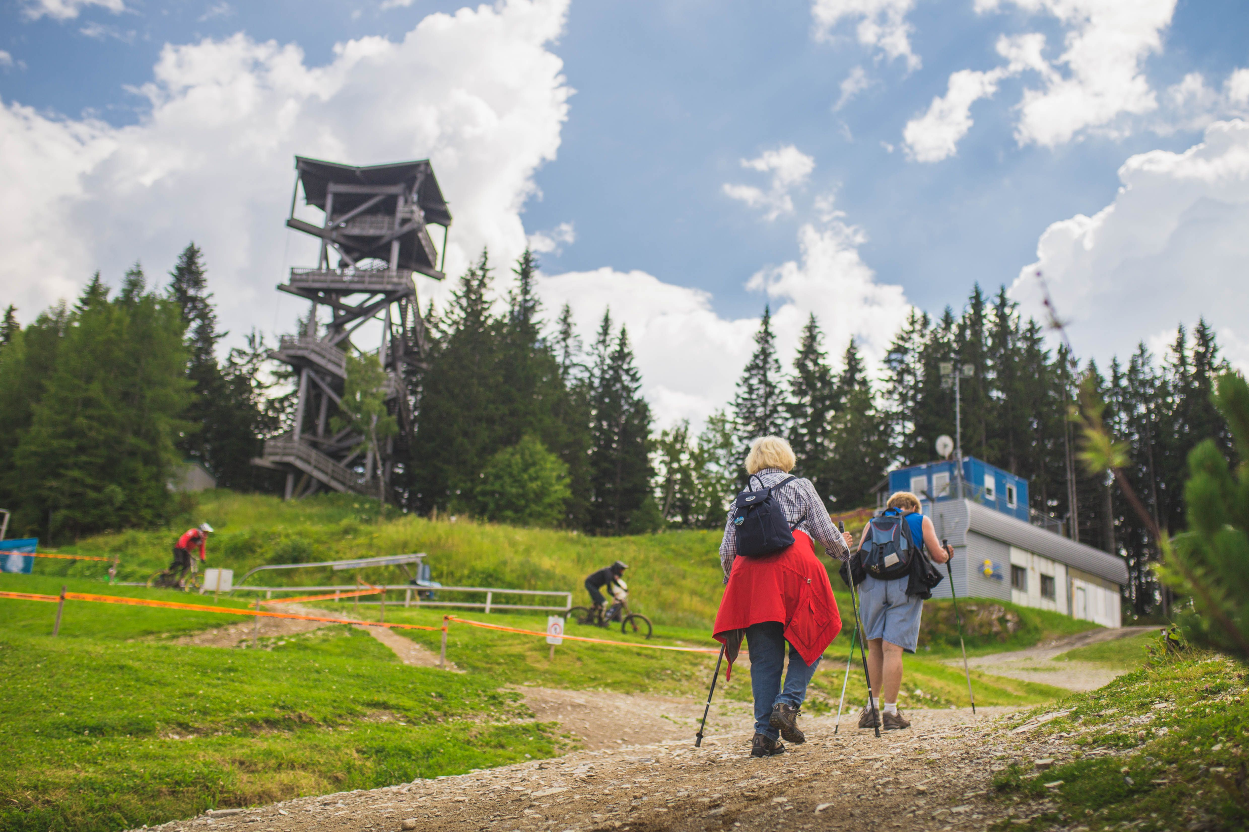 Zwei Wanderer gehen auf einem Weg bergauf, vorbei an einem Aussichtsturm und einem Gebäude, umgeben von Bäumen und einem klaren Himmel.