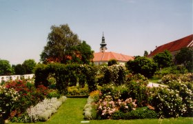 Historischer Garten mit Blumen und B&auml;umen, im Hintergrund ein Geb&auml;ude mit Turm.
