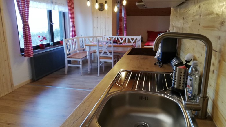 K&uuml;che in einem Dachgeschoss-Apartment mit Holzverkleidung, Sp&uuml;le, Esstisch und roten Vorh&auml;ngen.